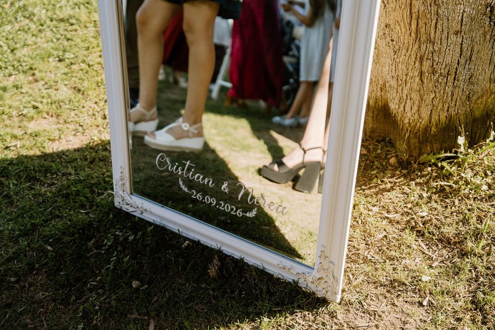White framed mirror on grass reflecting wedding guests' legs and shoes, with a personalized couple's names written in elegant script.