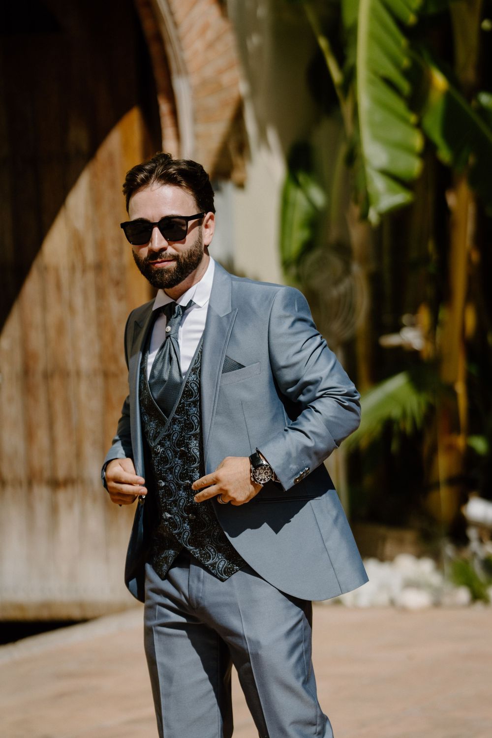 Bearded groom in sunglasses wearing a blue-gray suit with a patterned vest and tie, standing outdoors near tropical plants.