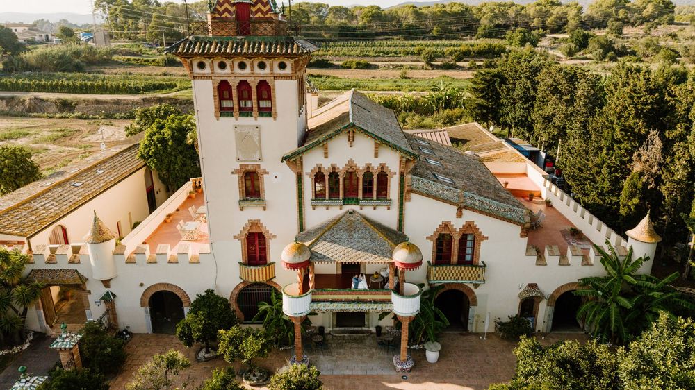 Aerial view of an elegant Mediterranean wedding venue with a tall tower, terracotta rooftops, a stone terrace, and lush green gardens.