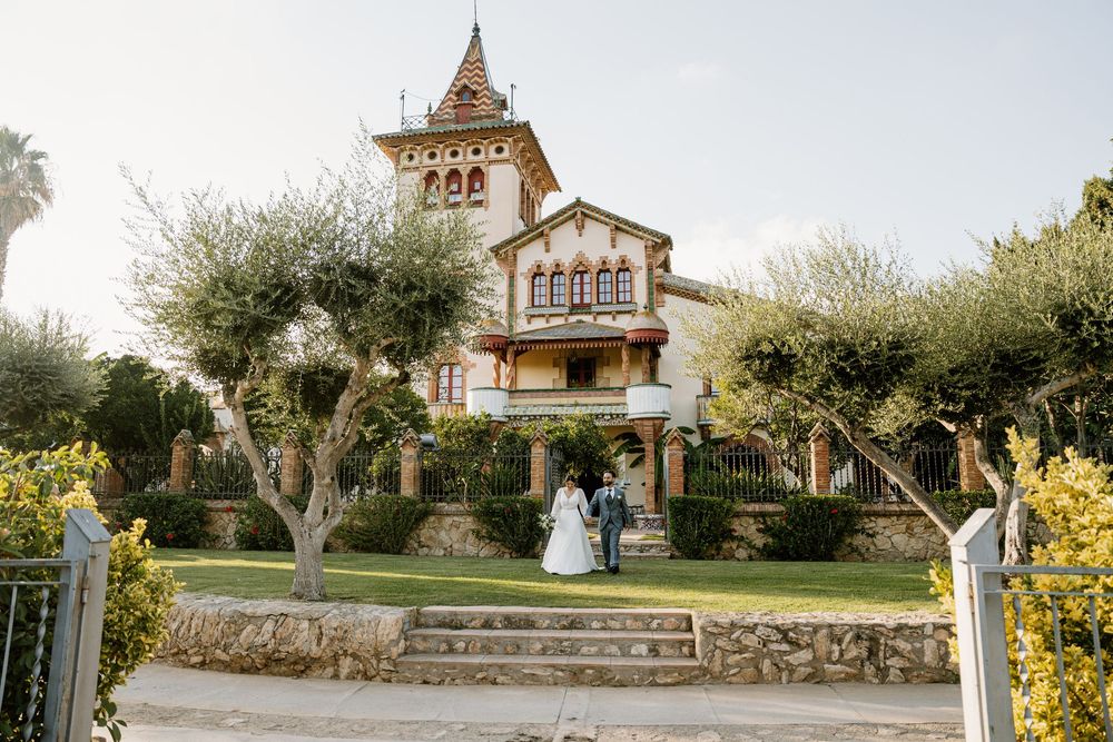 Bride and groom stand together on a green lawn in front of a historic villa with a tower, surrounded by olive trees on a sunny wedding day.
