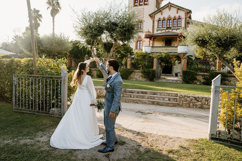 A bride in a white gown and a groom in a gray suit dance hand in hand on a gravel path outside a charming villa with gardens.
