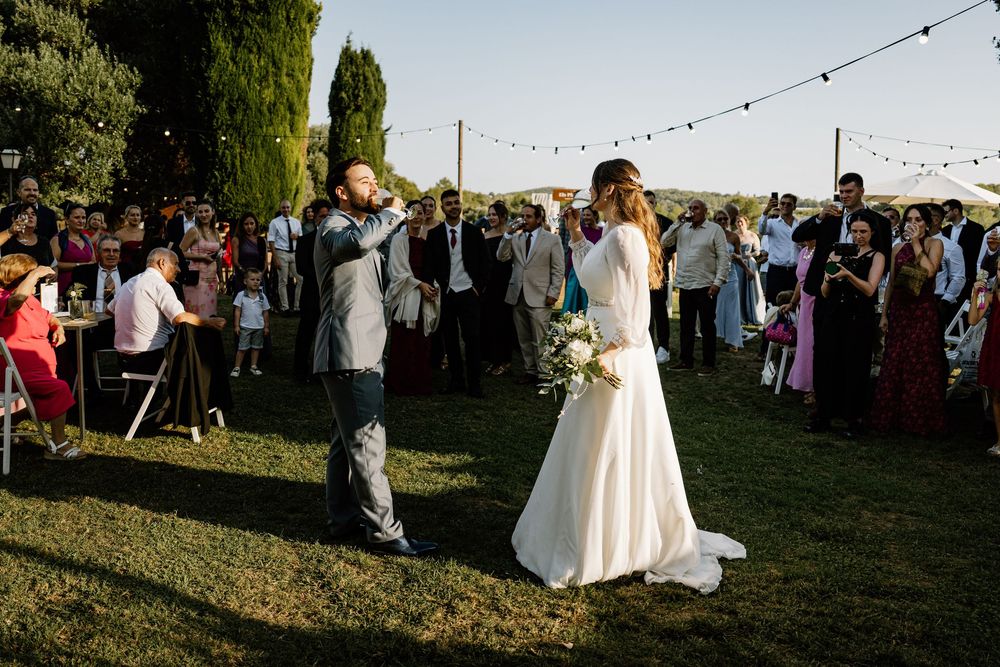 Bride in a white gown holding a bouquet stands facing the groom on the grass as wedding guests watch at an outdoor garden ceremony.