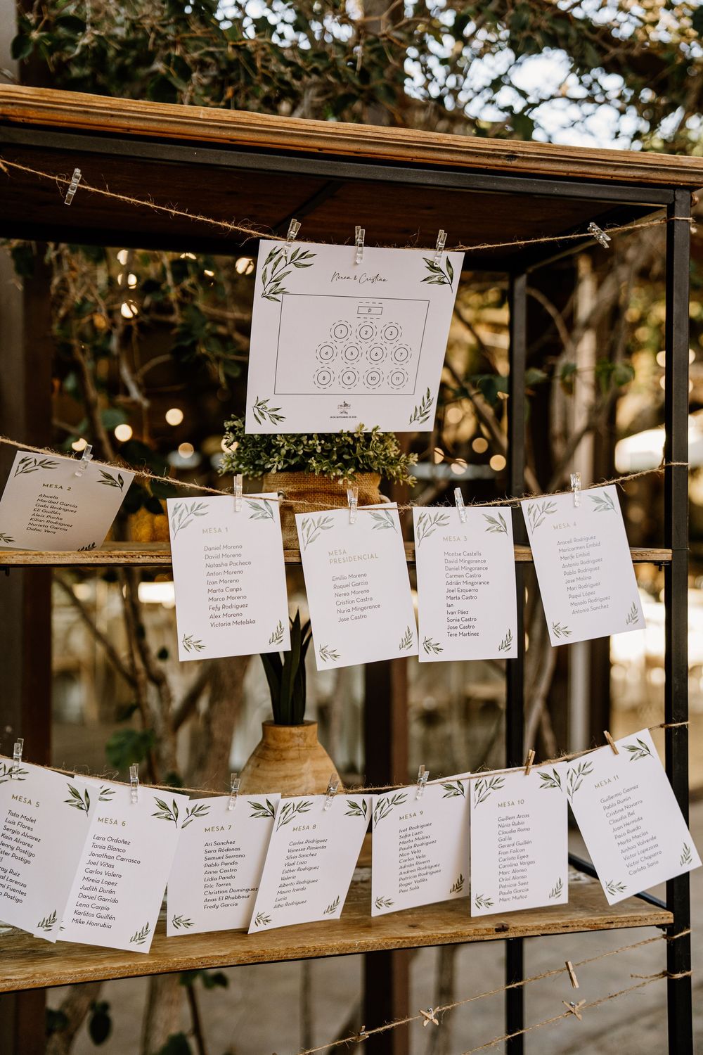 Rustic wedding seating chart with a table layout and guest list cards pinned on twine across wooden shelves with greenery and string lights.