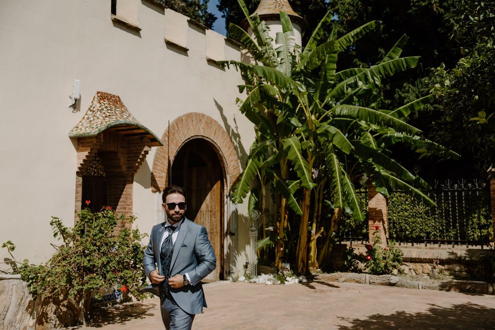 Man in sunglasses and a gray suit stands in a sunlit courtyard beside a rustic arched doorway and tall green banana plants.
