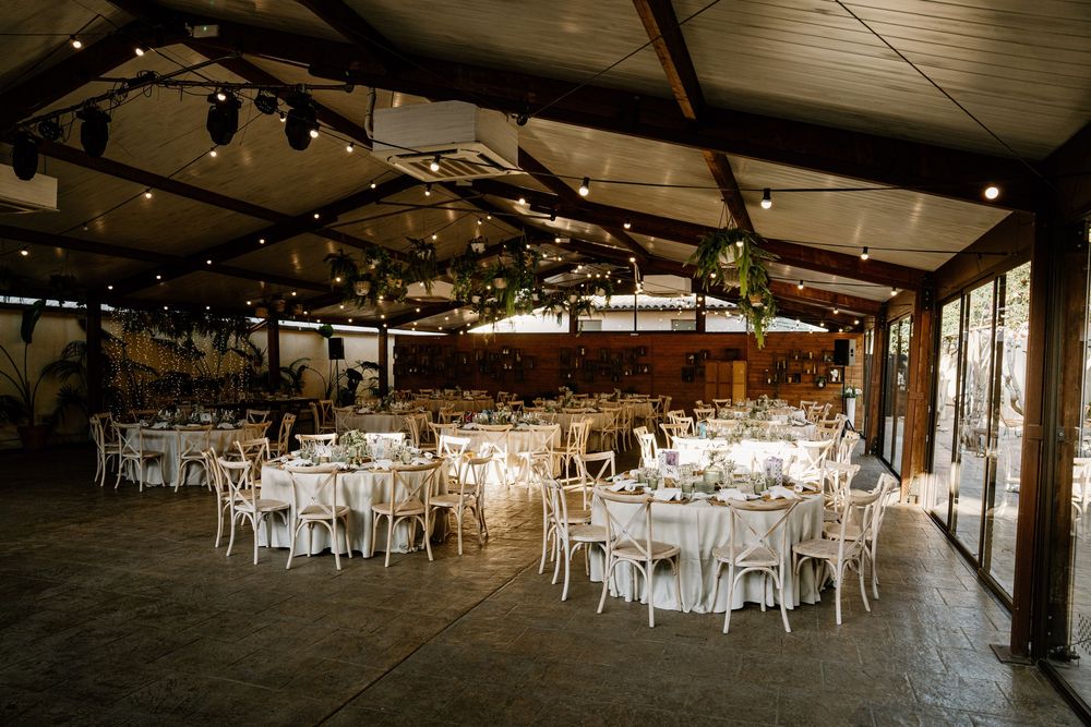 Rustic indoor wedding reception venue with round tables, white chairs, hanging greenery and string lights under a wooden beamed ceiling.