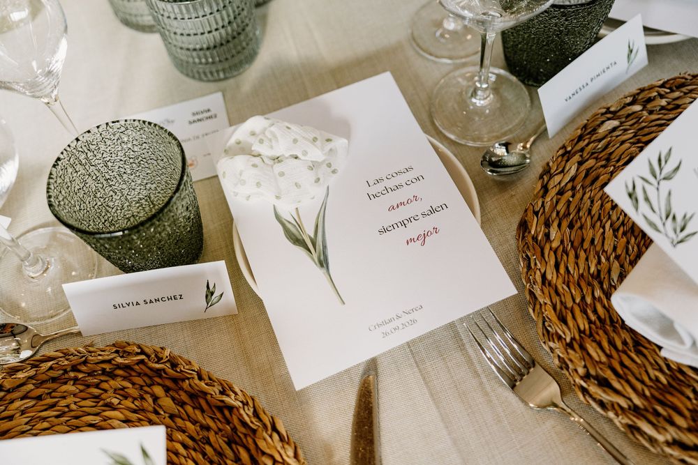 Elegant wedding table setting with a botanical menu card, woven rattan chargers, green glass tumblers, name cards, and silverware on linen.