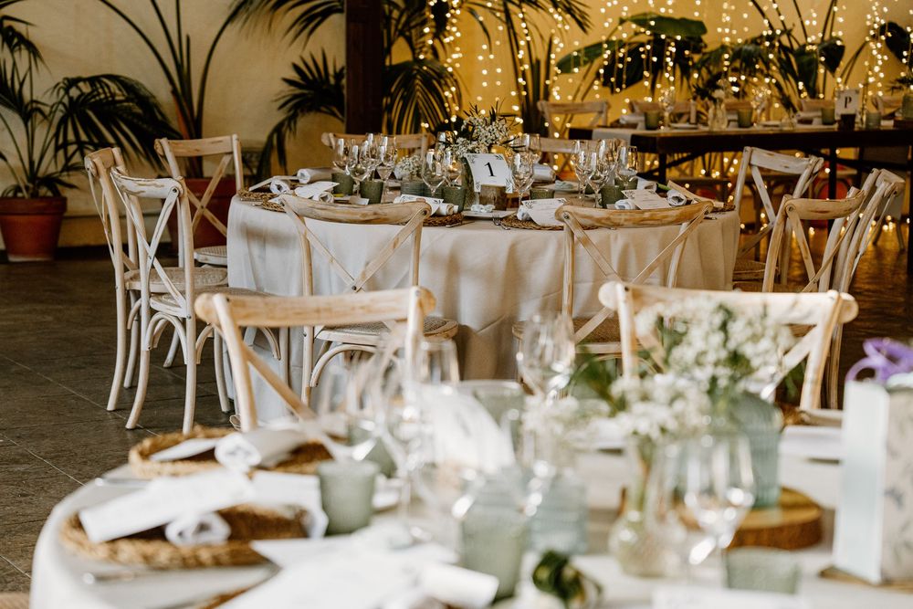 Elegant wedding reception tables with white linens, wooden cross-back chairs, glassware, greenery centerpieces and warm string lights.