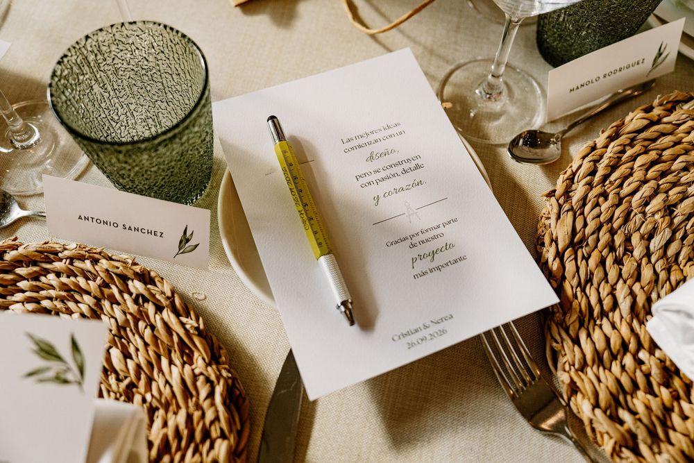 Elegant wedding table setting with a printed menu card, yellow pen, woven rattan placemat, textured green glass and a guest name place card.