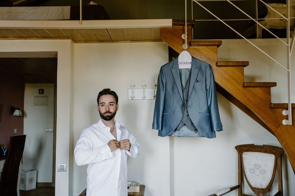 Groom in a white dress shirt buttoning his cuffs beside a blue suit jacket hanging on a wall near a wooden staircase.