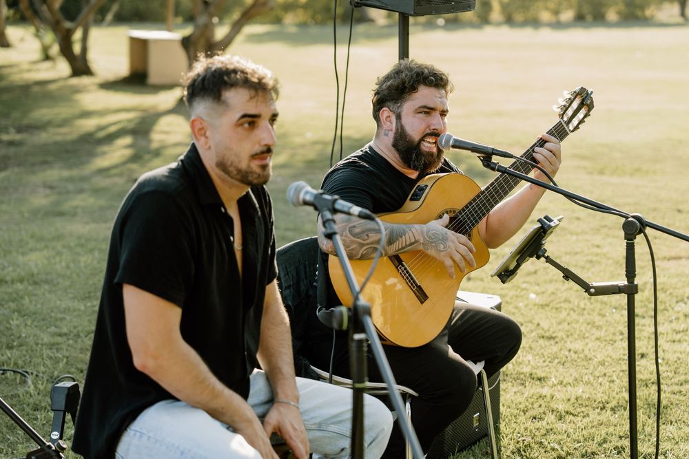 Two musicians perform at an outdoor wedding ceremony, one playing an acoustic guitar while both sit near microphones on a sunlit lawn.