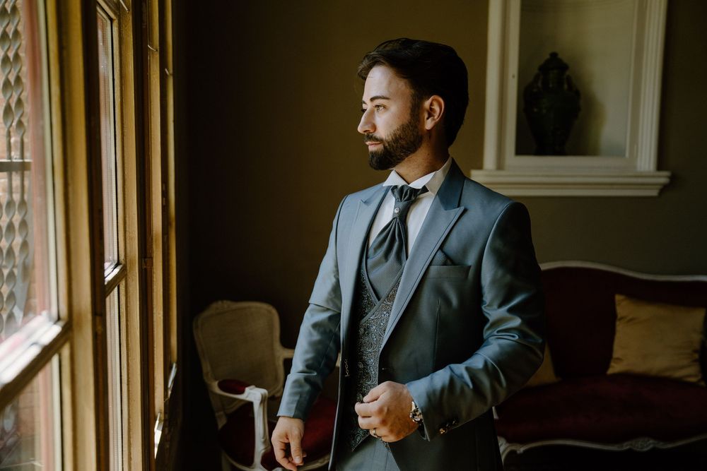 Bearded groom in a gray three-piece suit and patterned tie adjusts his cuff while gazing out a sunlit window in a cozy vintage room.
