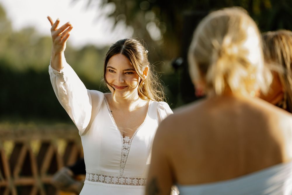 A smiling bride in a white lace-trimmed dress raises her hand in celebration during an outdoor wedding ceremony as guests look on.