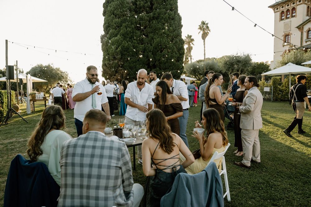 Guests mingle and chat at an outdoor garden party on a green lawn under string lights, with tents and tall trees in the background.