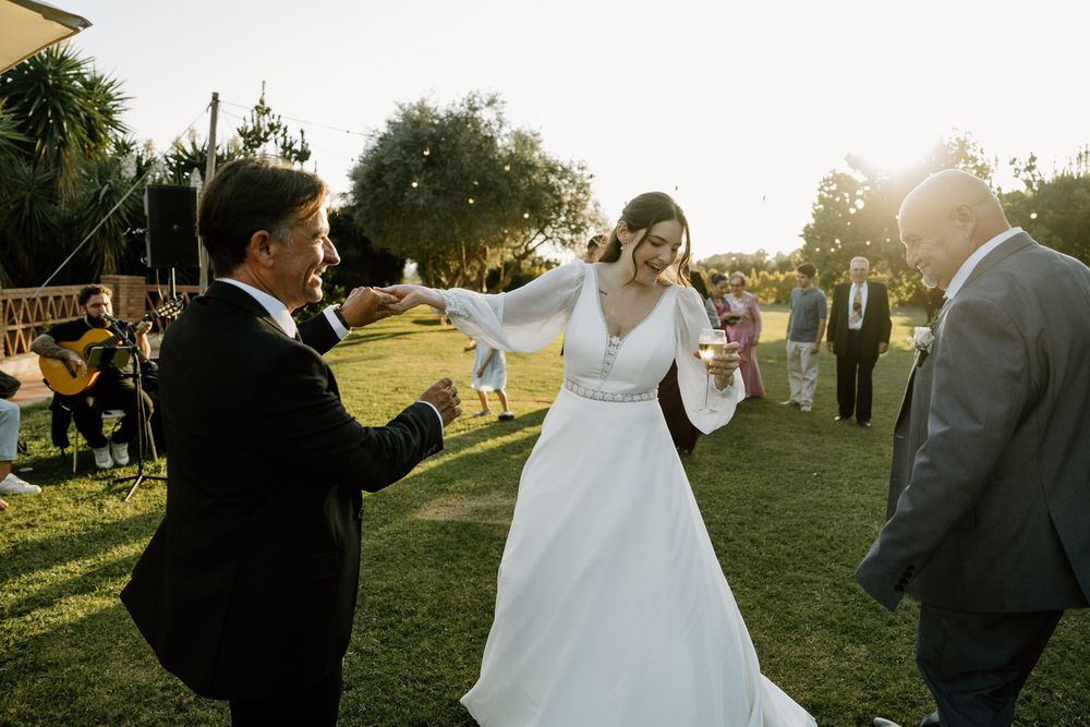 Bride in a flowing white gown dances joyfully with a groom in a dark suit on a sunlit lawn while guests and a guitarist look on.