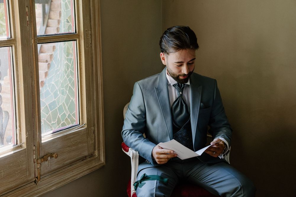 Groom in a gray suit and tie sits by a window reading a letter on his wedding day.