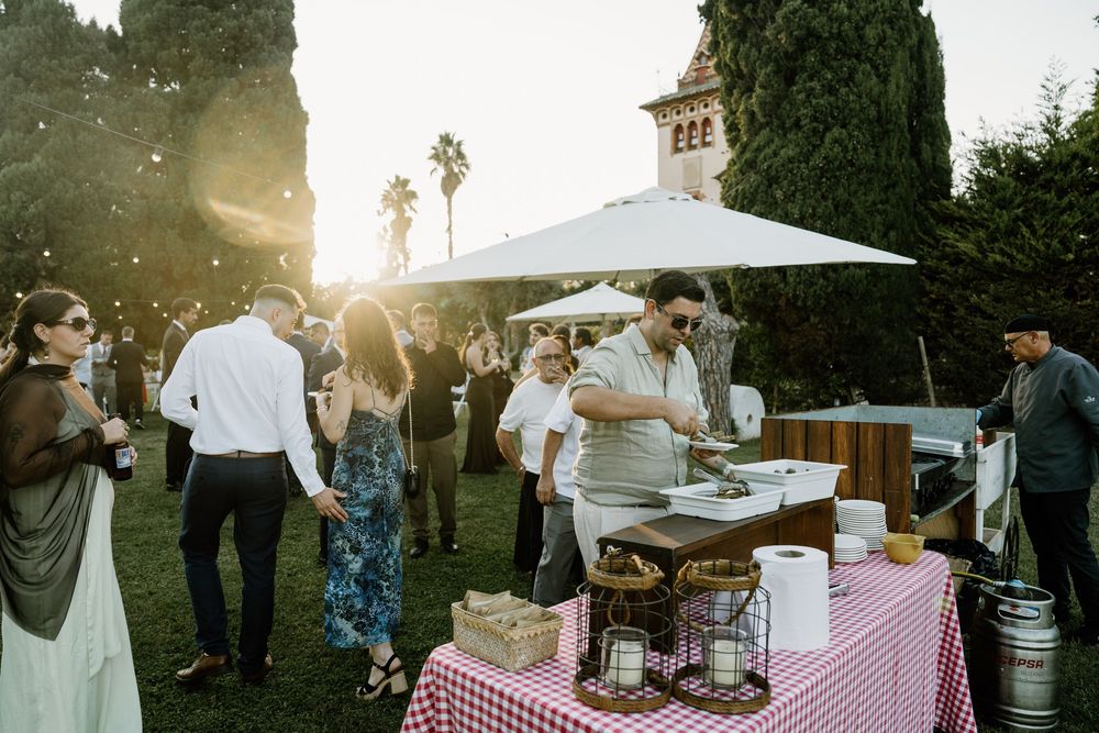 Guests mingle at an outdoor evening reception on a lawn, with a man serving food at a checkered buffet table under a white umbrella.