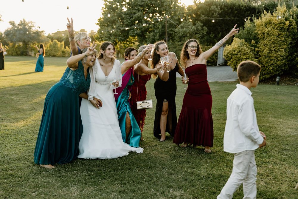 Bride in a white gown and bridesmaids in green and burgundy dresses reach upward on a lawn as a young boy in white watches.