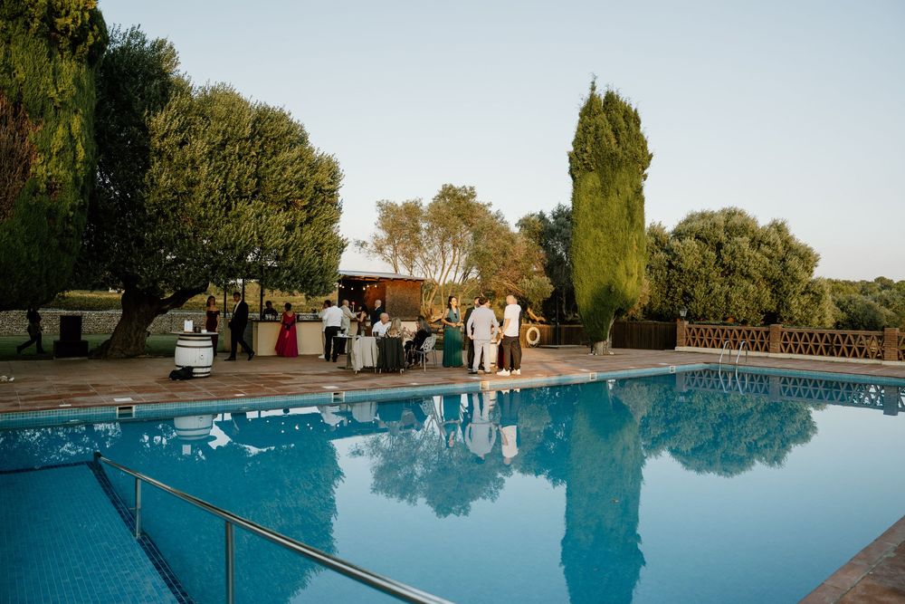 Guests gather on a stone terrace beside a calm outdoor swimming pool at sunset, surrounded by tall cypress and green trees.