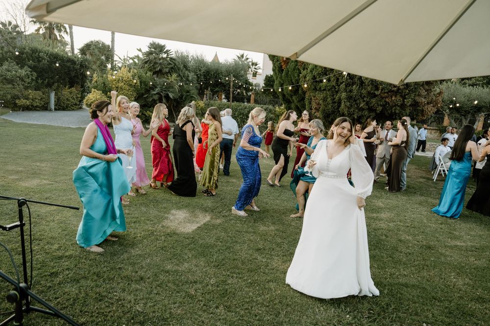 Bride in a white gown stands on a lush green lawn as colorful dressed wedding guests dance together at an outdoor garden celebration.