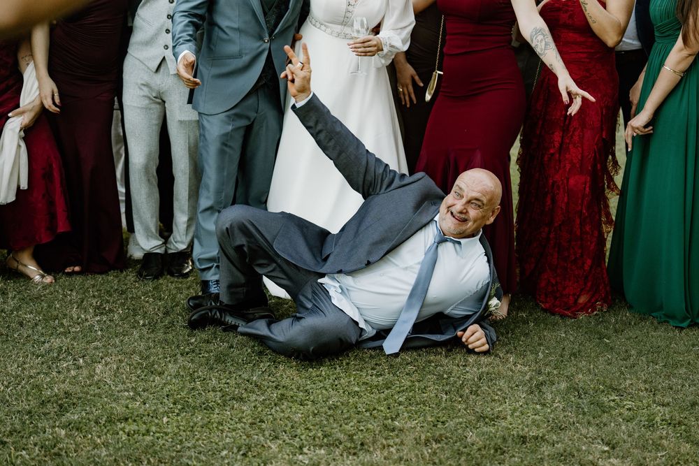 Smiling bald man in a gray suit and blue tie sprawls playfully on the grass as wedding guests in colorful dresses stand behind him.