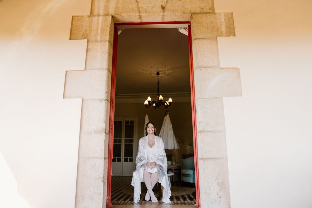 Bride in a white lace robe sits in a stone doorway with a chandelier and wedding dress hanging in the elegant room behind her.