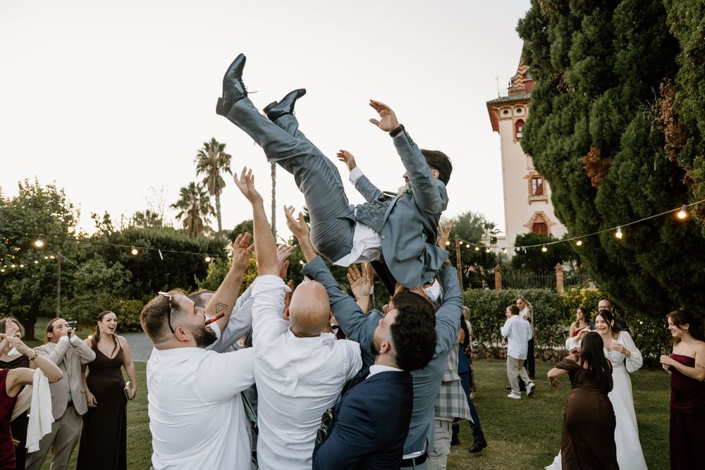 Wedding guests toss a groom in a gray suit into the air outdoors at a festive garden reception with string lights and palm trees.
