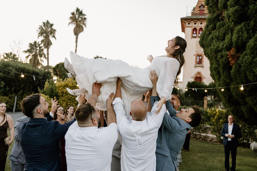 Wedding guests lift a bride in a white gown into the air at an outdoor garden celebration with palm trees and a historic tower behind them.