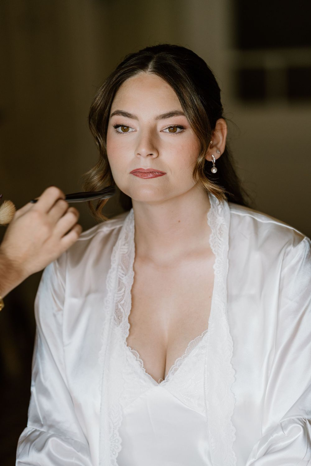 Bride in a white satin robe with lace trim and pearl earrings having her makeup applied by an artist while getting ready for her wedding.