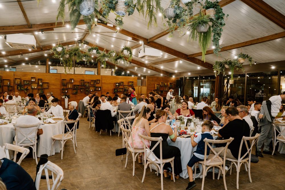 Wedding guests seated at round tables enjoy a reception in a rustic hall with wood beams and hanging greenery garlands.