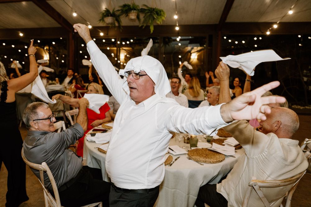 Guests at a festive banquet dinner wave white napkins in the air while a man with a napkin on his head dances beside a table.