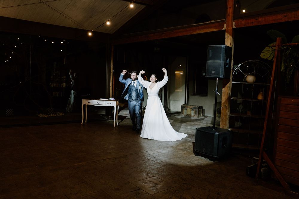 Bride in a white gown and groom in a blue suit raise their arms in celebration on a dimly lit wedding reception dance floor.