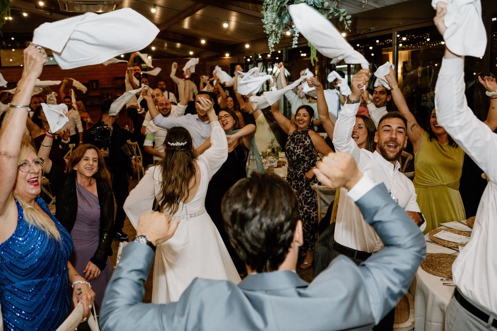 Wedding guests cheer and wave white napkins in the air while dancing together on a lively reception dance floor.