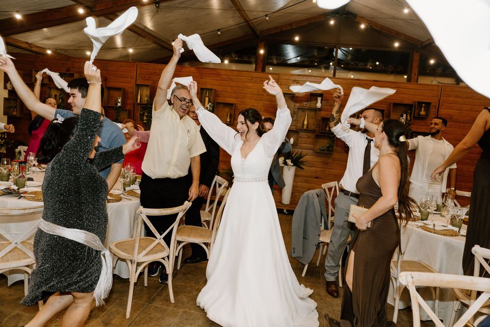 Wedding guests wave white napkins in the air while a bride in a flowing white gown dances in the center of a rustic reception hall.
