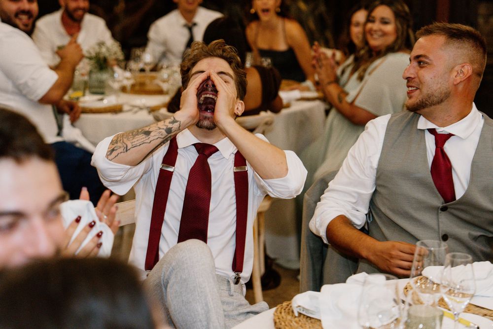 Groomsman in a white shirt, burgundy tie and suspenders cups his hands to shout while seated wedding guests laugh at a reception table.