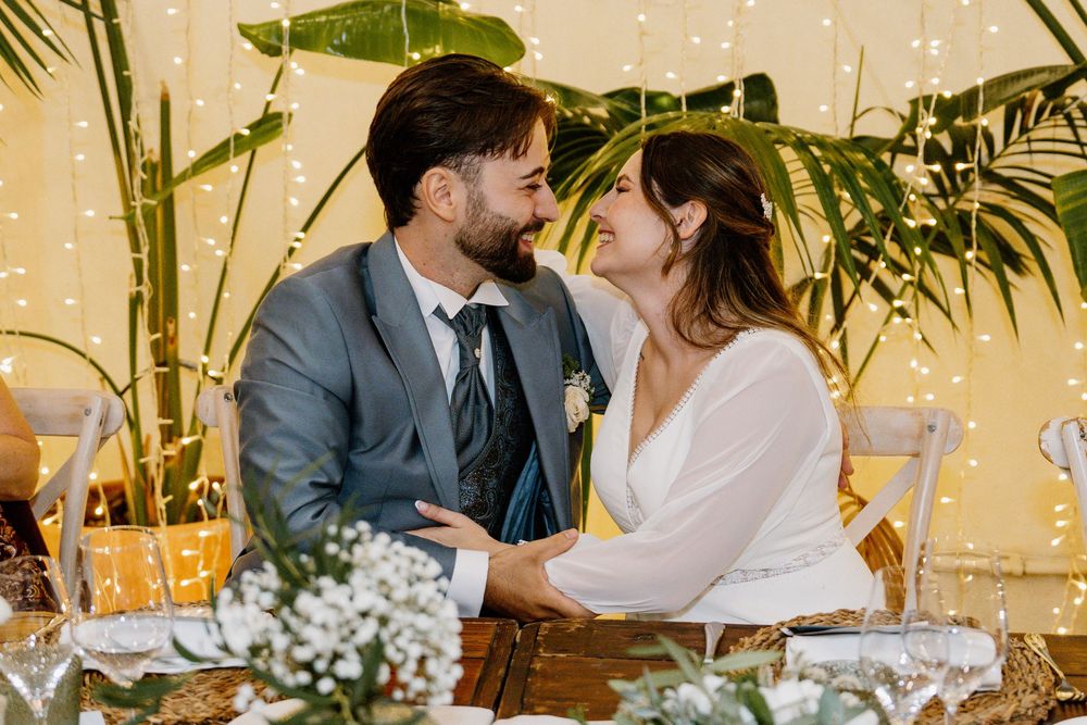 Smiling bride in a white dress and groom in a gray suit share a tender moment at a wedding reception table with flowers and string lights.