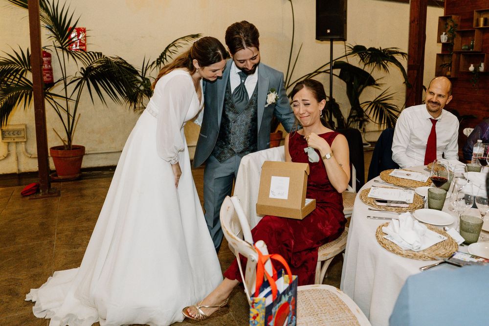 Bride in a white gown and groom in a gray suit hand a gift box to a smiling woman in a red dress seated at a wedding reception table.