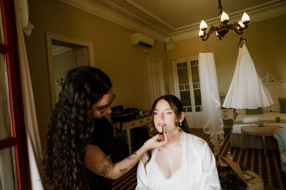 Makeup artist applies lipstick to a bride in a white robe in an elegant room with a chandelier and a wedding dress hanging nearby.