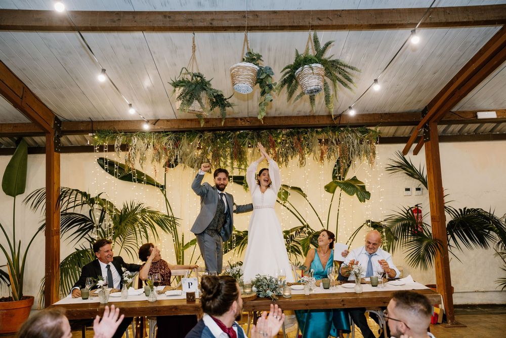 Bride and groom raise their arms in celebration at a wedding reception beneath hanging plants while guests cheer at a long wooden table.
