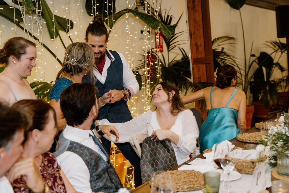 Smiling wedding guests gather around a reception table as a man in a vest stands and speaks, with string lights and plants behind them.