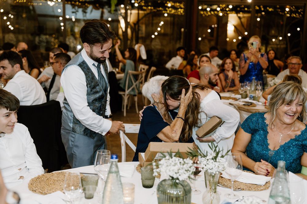 Emotional bride leans on a table covering her face while the groom and wedding guests look on at a candlelit reception with string lights.