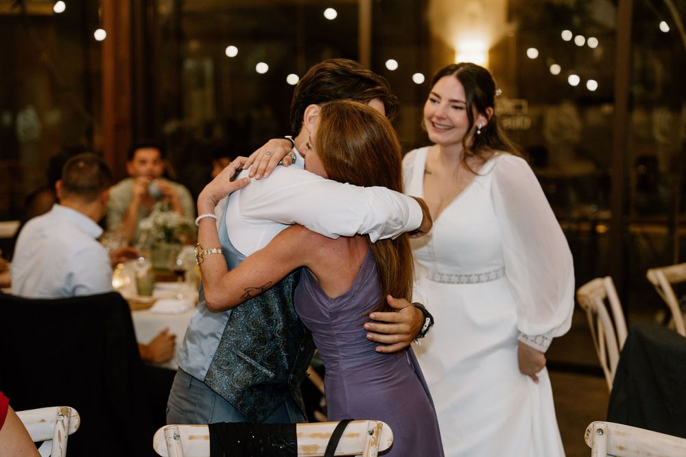A guest in a purple dress hugs a man in a denim vest at a wedding reception while a bride in a white gown smiles nearby.