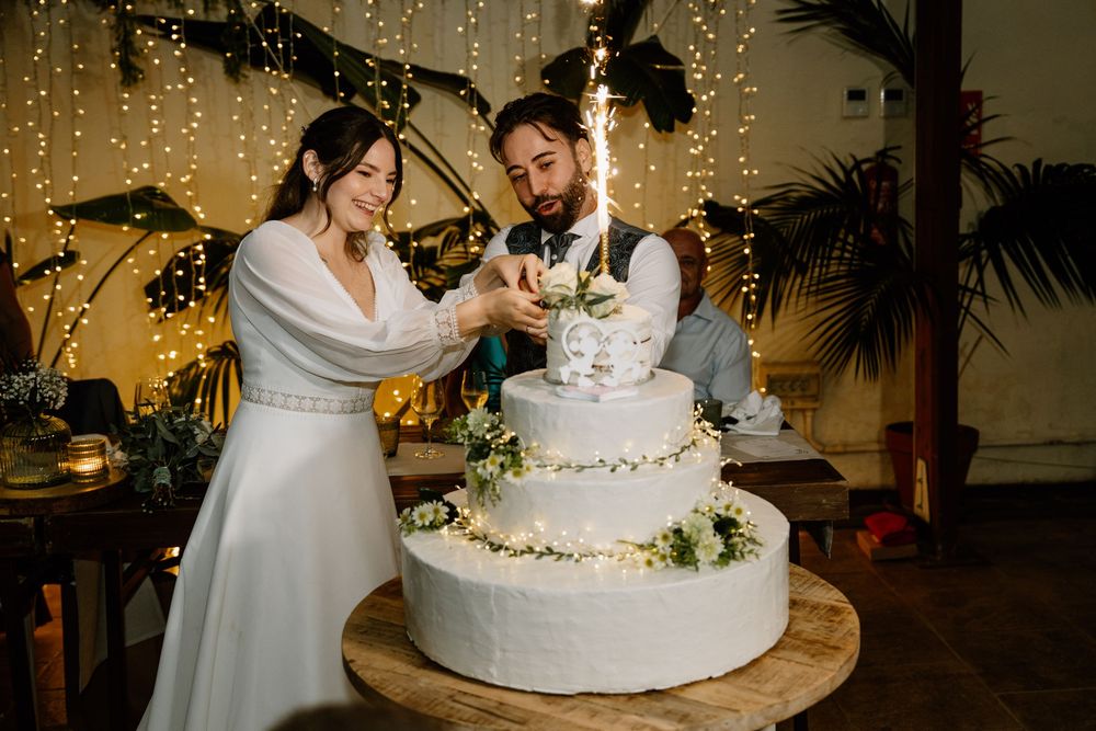Bride and groom cutting a white tiered wedding cake decorated with greenery and string lights at an intimate indoor wedding reception.