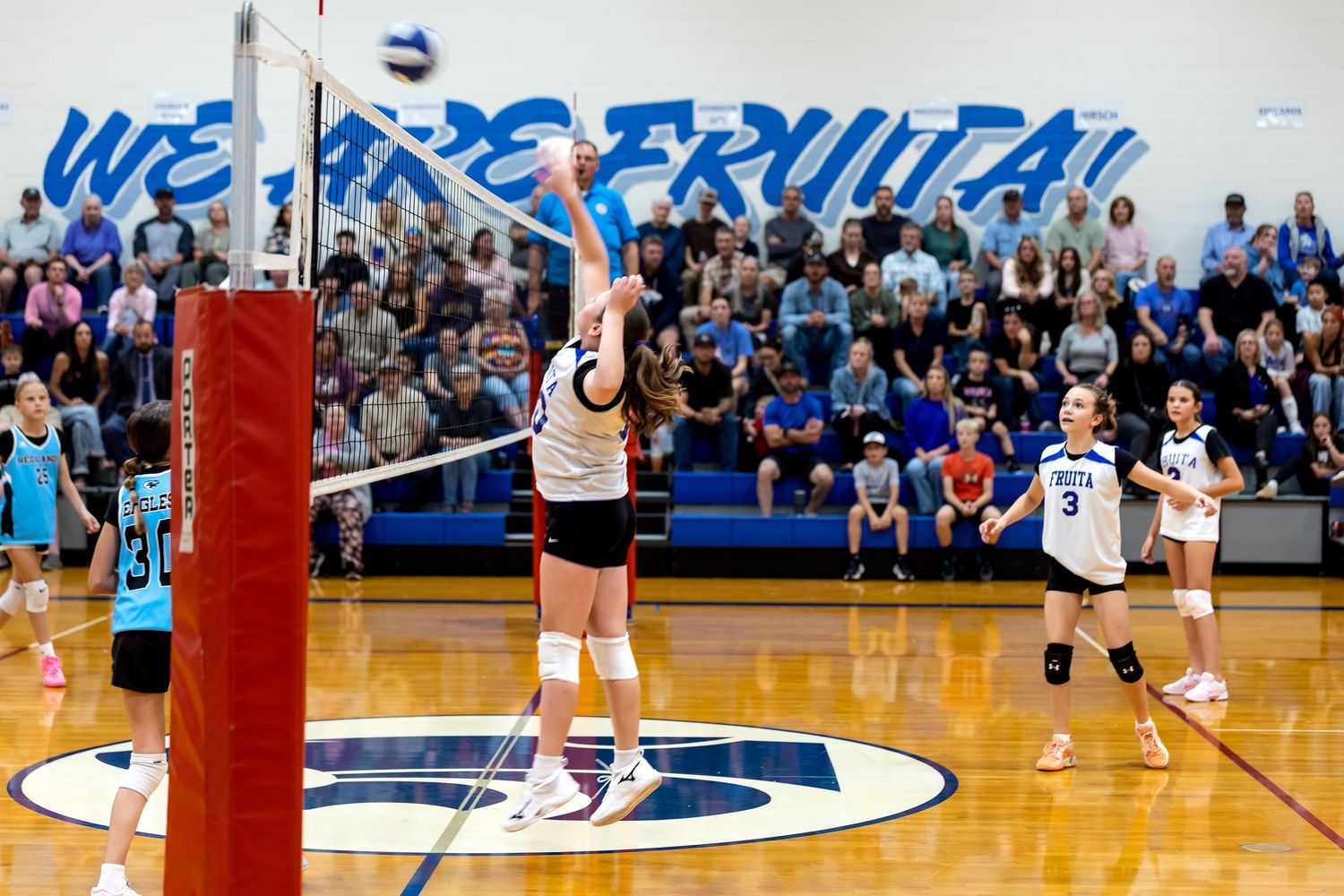 A volleyball player jumps at the net to spike the ball during a youth match with fans watching from the stands behind her.