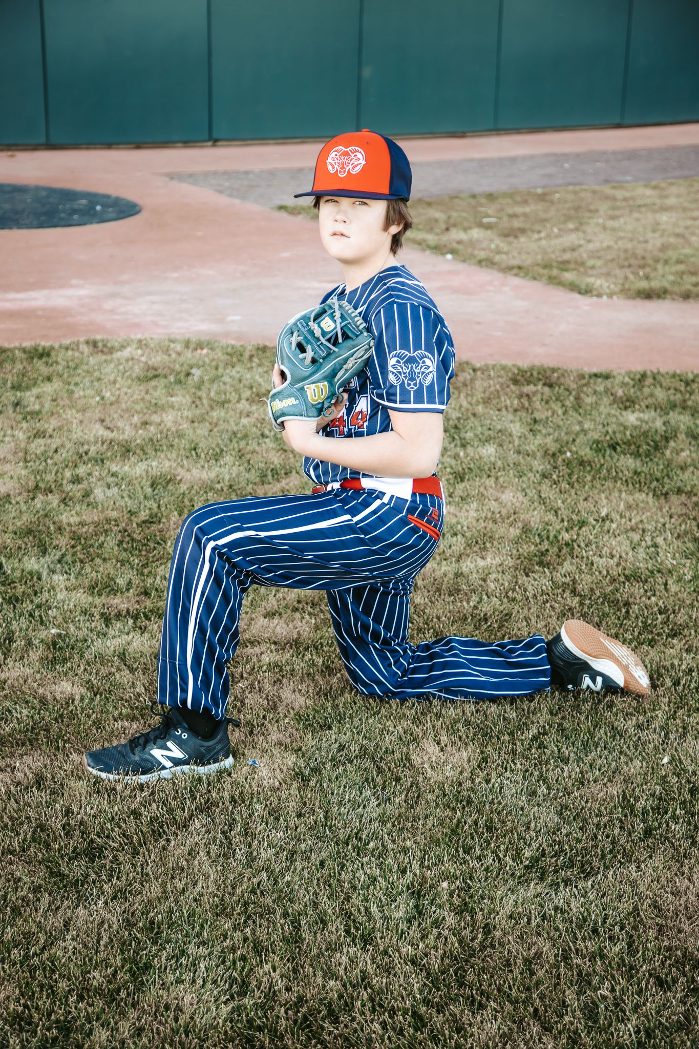 A young baseball player in a navy pinstripe uniform and orange cap kneels on the grass holding a glove in a ballpark.