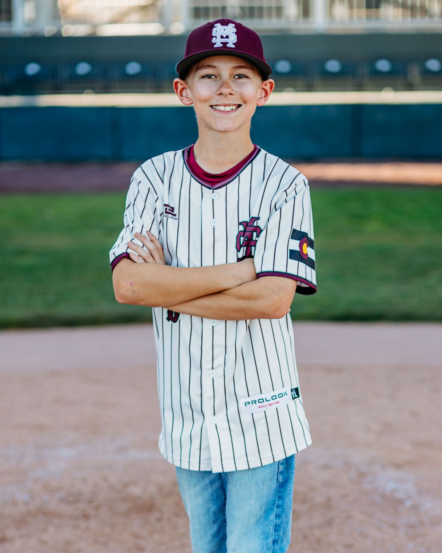 Smiling boy in a maroon cap and pinstripe baseball jersey with a Colorado flag patch stands with arms crossed on a baseball field.