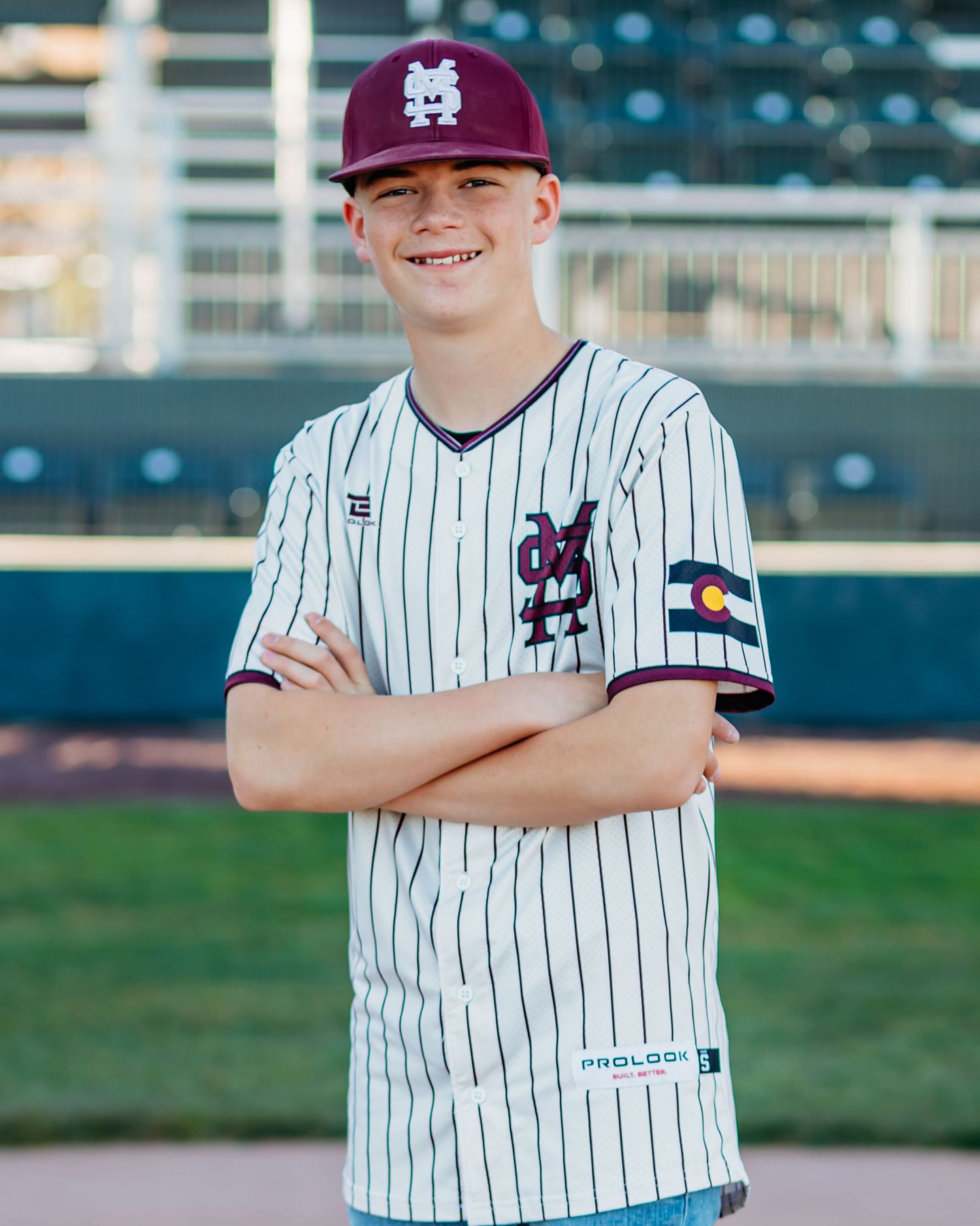 Grinning teen baseball player in a maroon cap and white pinstripe jersey with a Colorado flag patch stands arms crossed at a ballpark.