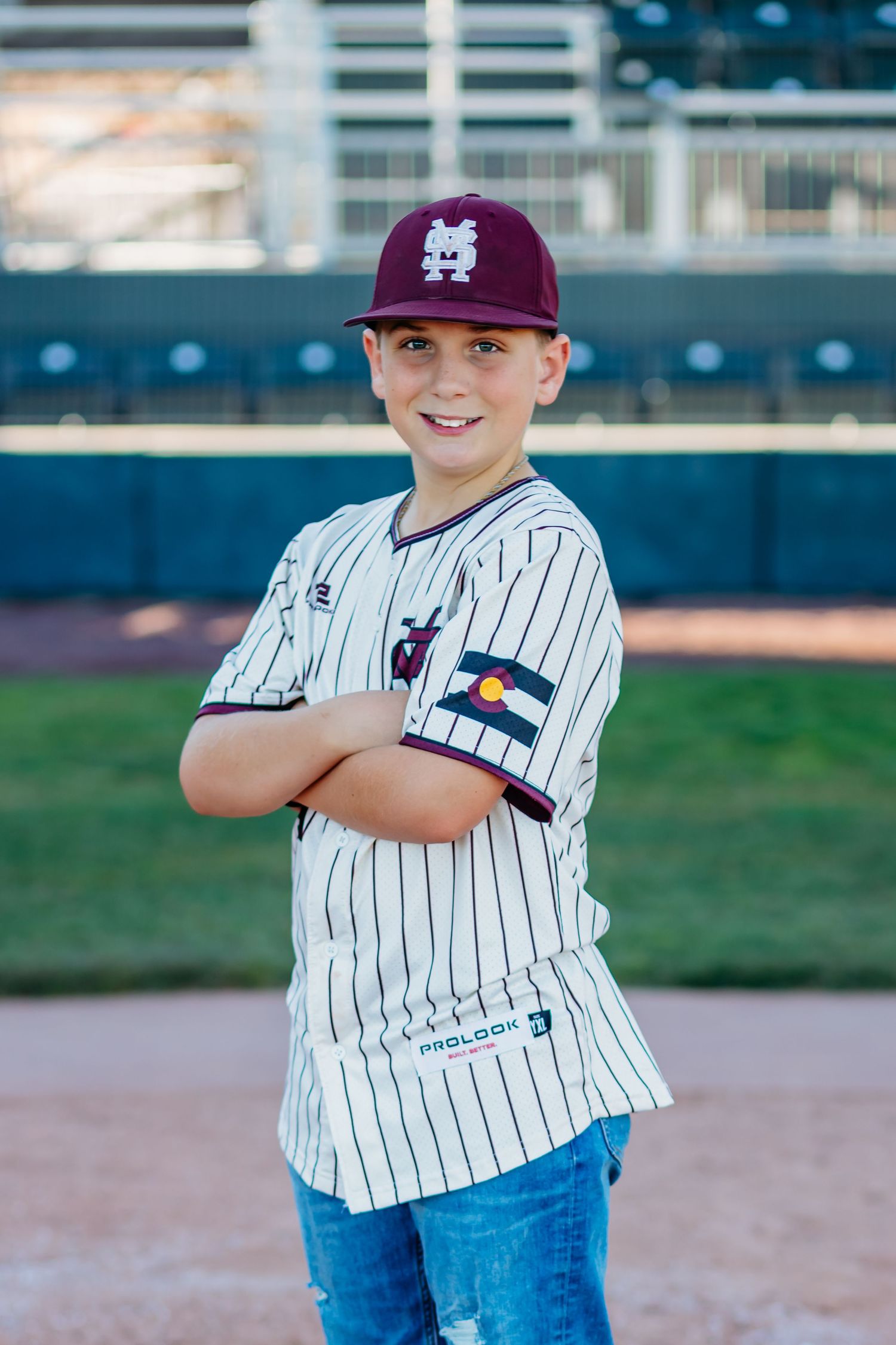 Smiling youth baseball player in a maroon cap and pinstripe jersey stands with arms crossed on the field.