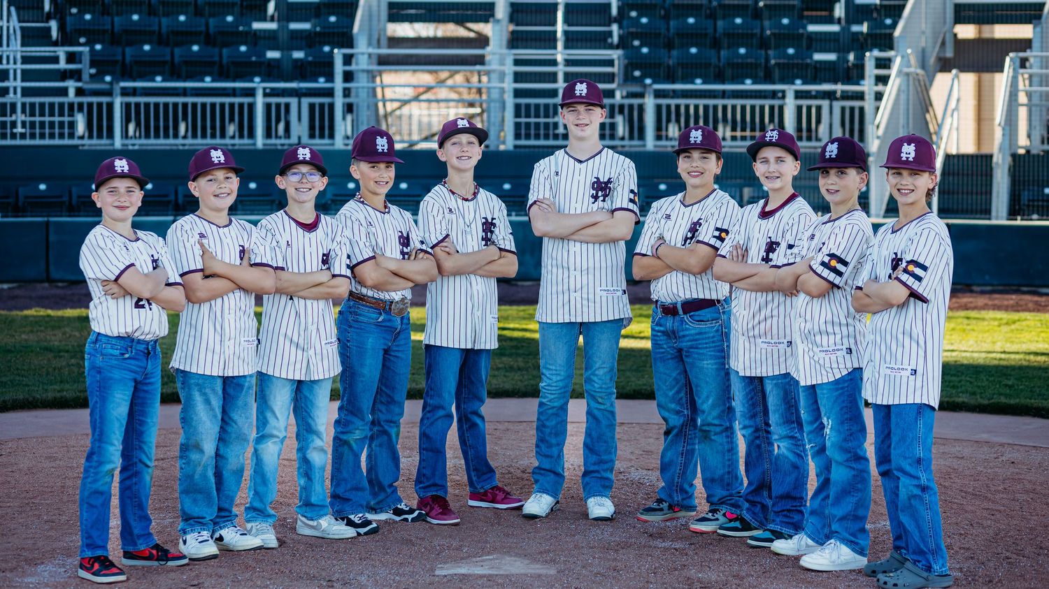Youth baseball team in maroon caps and pinstripe jerseys poses with arms crossed on the infield dirt.