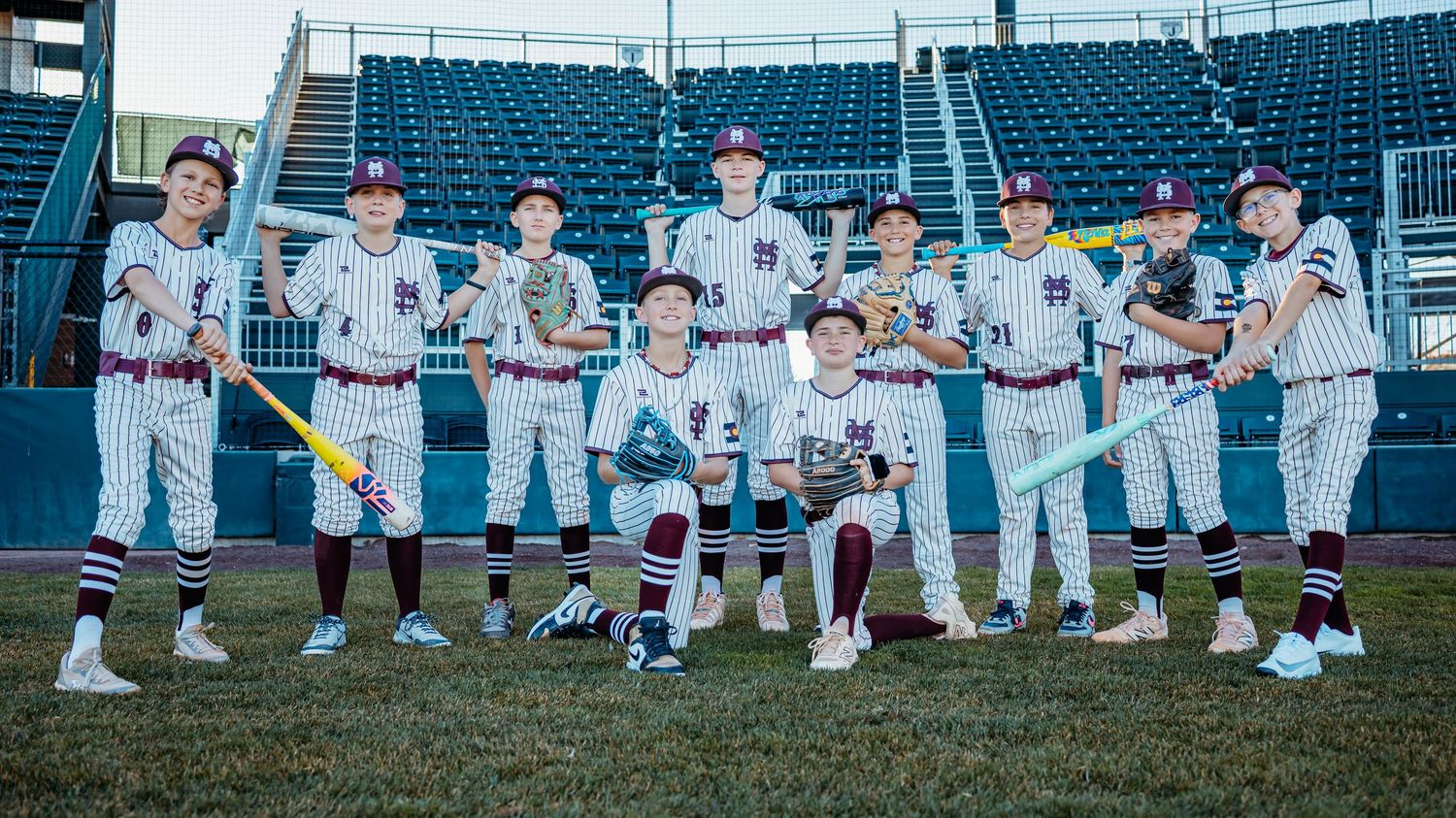 Youth baseball team in pinstripe uniforms poses with bats and gloves on the infield in front of stadium seating.