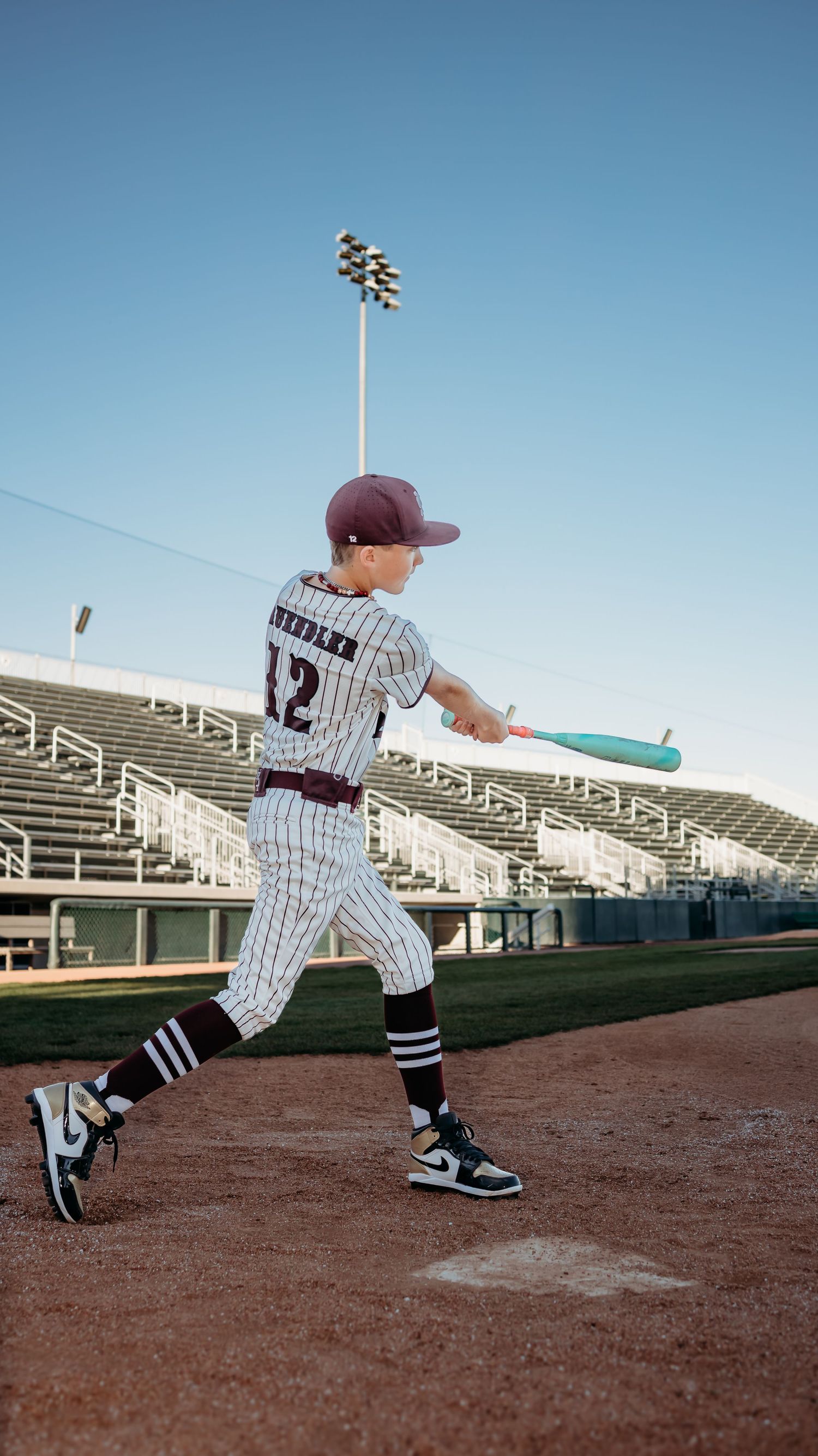 Young baseball player in a pinstripe uniform and maroon cap swings a teal bat at an empty stadium.